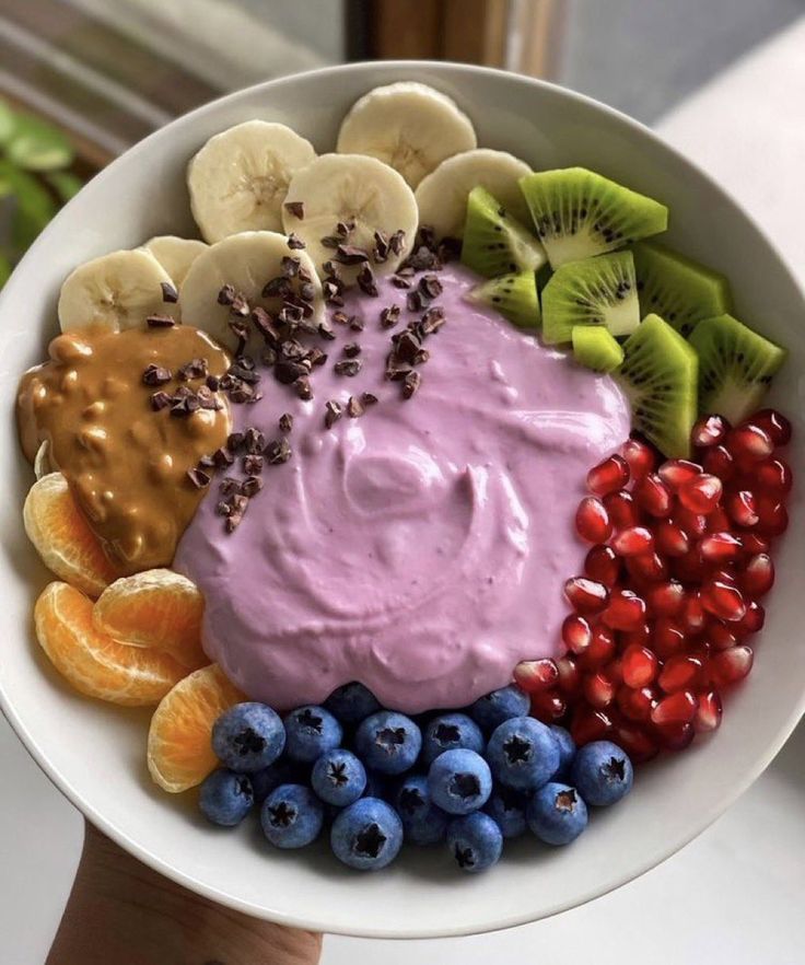 a white bowl filled with fruit and dip surrounded by kiwis, oranges, blueberries