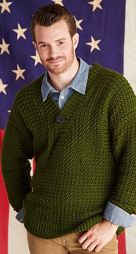 a man standing in front of an american flag wearing a green sweater and tan pants