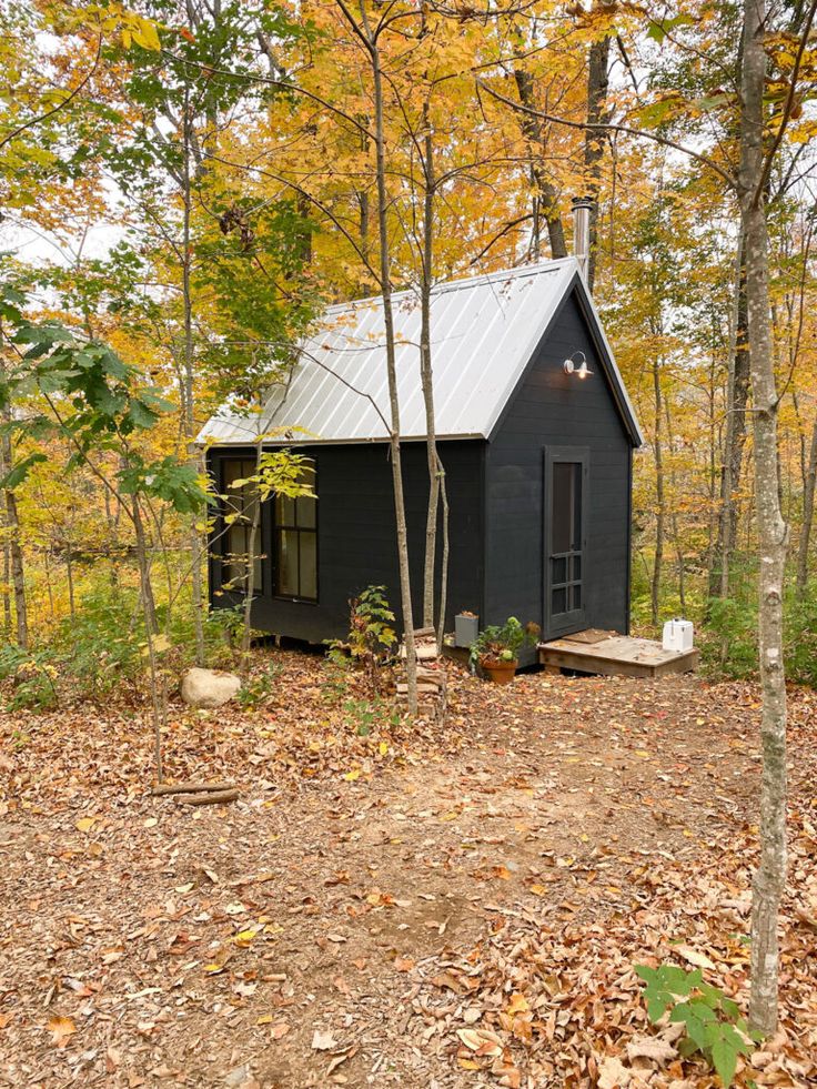 a small black cabin in the woods surrounded by trees and leaves with fall foliage around it