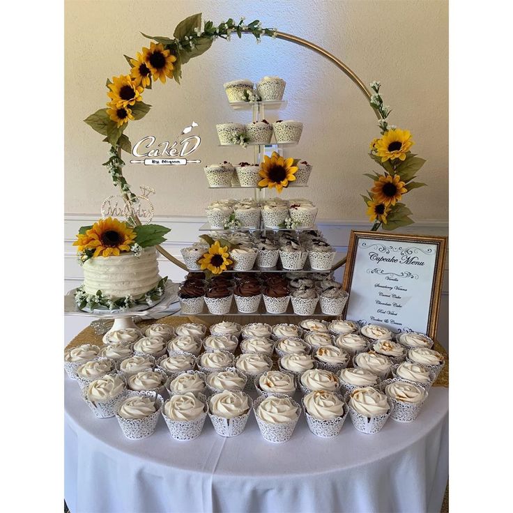 a table topped with lots of cupcakes next to a cake and sunflower