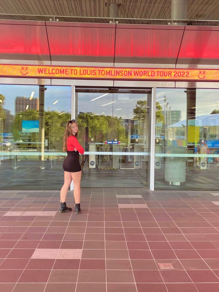 a woman standing in front of a building with her back to the camera, wearing short black shorts and a red shirt