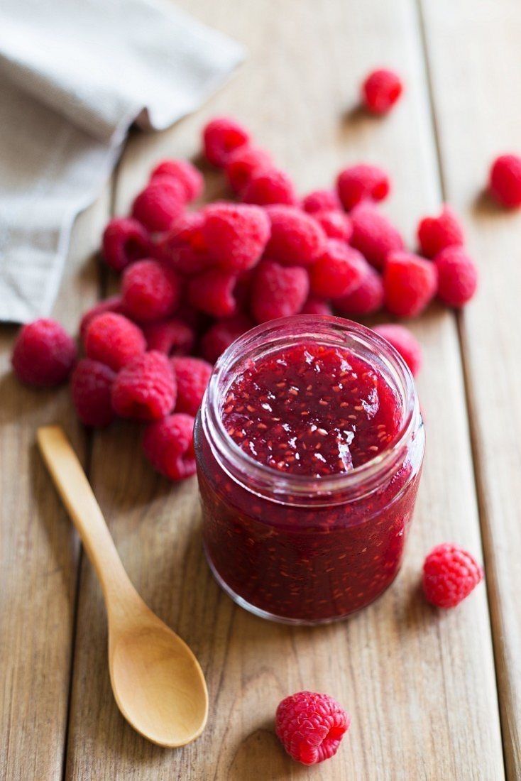 raspberry jam in a glass jar with spoon on wooden table next to fresh raspberries