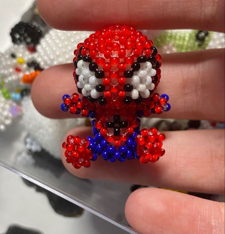 a hand holding a red, white and blue beaded spiderman brooch on it's finger