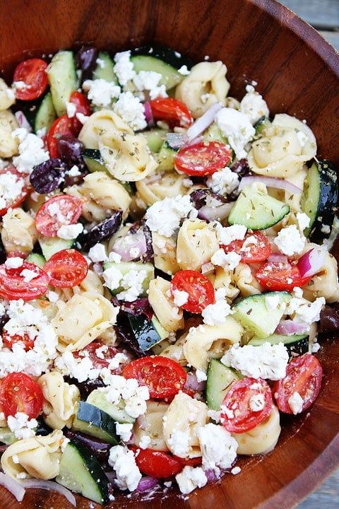 a wooden bowl filled with pasta salad and fettuccine, tomatoes, cucumbers, and cheese