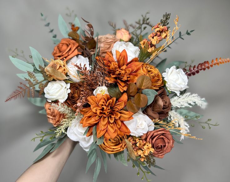 a bridal bouquet with orange, white and brown flowers is held by someone's hand