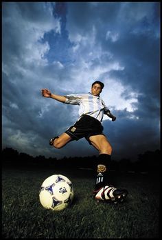 a man kicking a soccer ball on top of a lush green field with clouds in the background