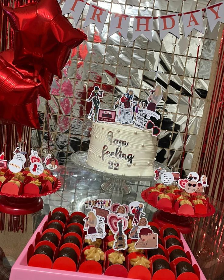 a birthday cake and cupcakes on display in front of a party backdrop with red balloons