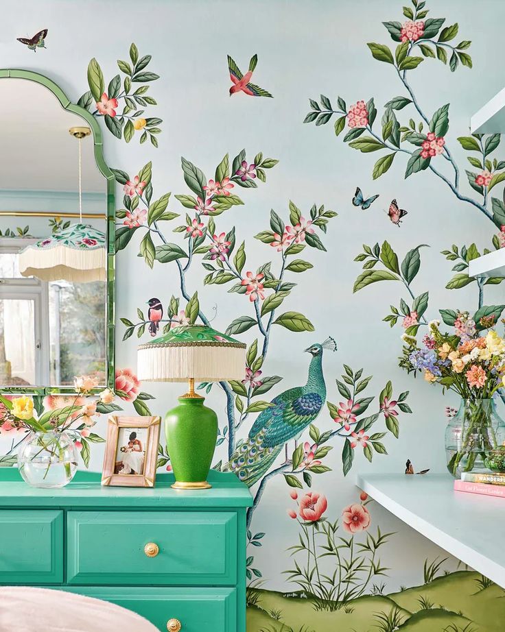 a room with flowers and birds painted on the wall next to a dresser, mirror and lamp