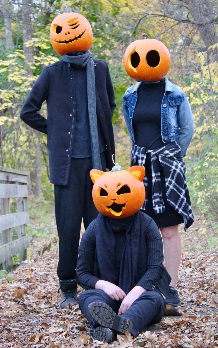 three people wearing pumpkin heads in the leaves with one person sitting on the ground next to them