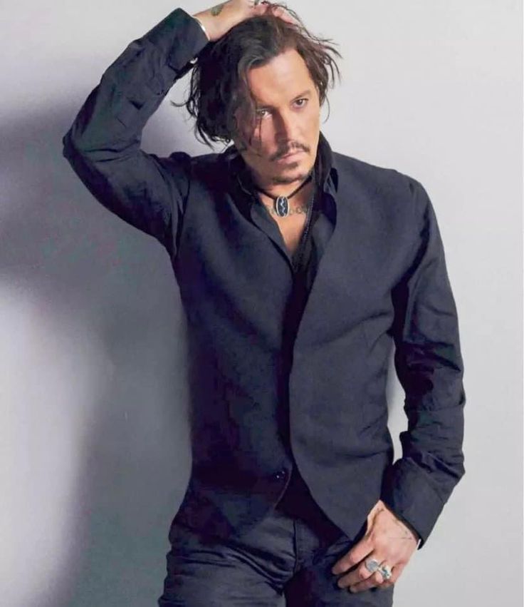 a man with long hair wearing a black shirt and jeans leaning against a wall holding his hand on his head