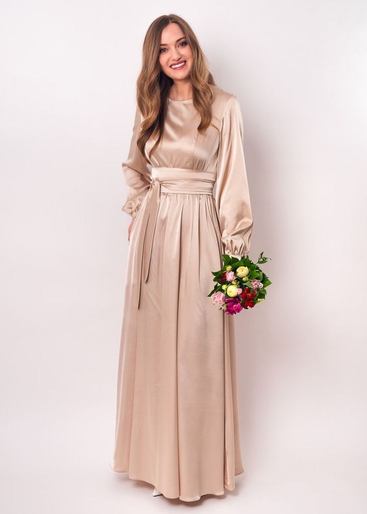 a woman in a long beige dress holding a flower bouquet and smiling at the camera