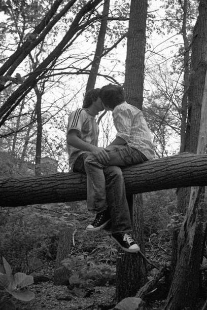 two people are sitting on a log in the woods, one is kissing the other