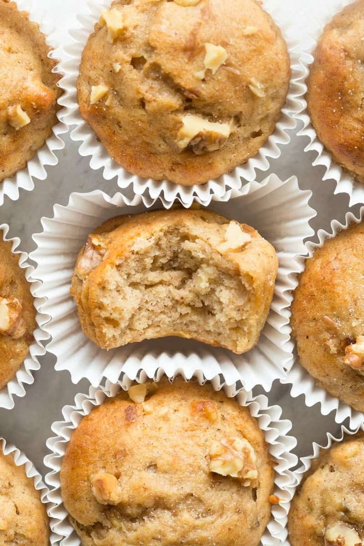 twelve banana muffins in white paper cups on a counter top with one cut in half