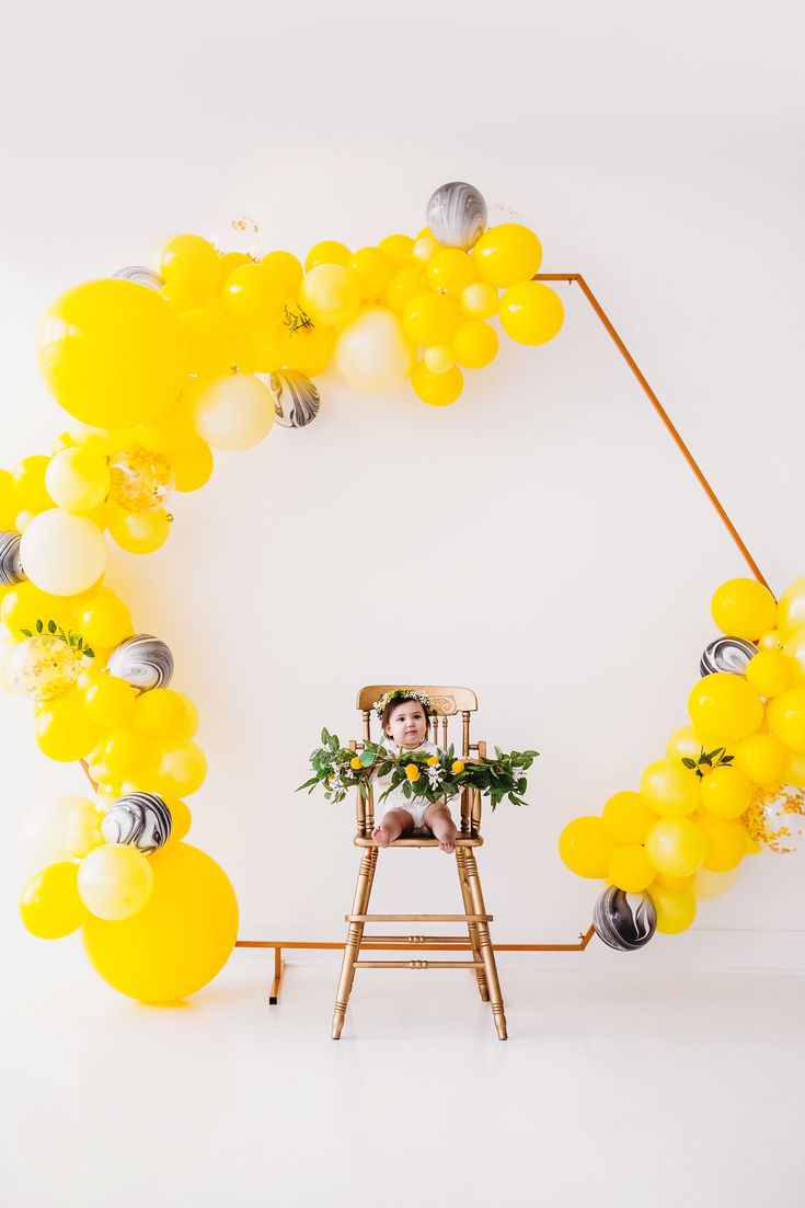 a man sitting in a chair surrounded by yellow balloons and greenery on a white background