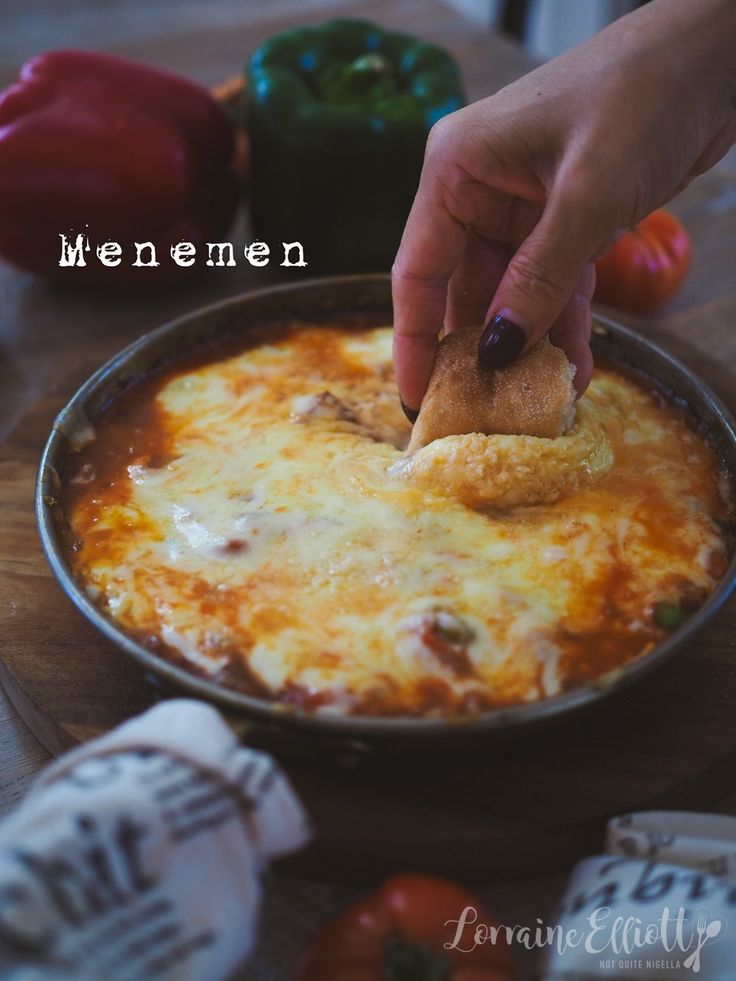 someone dipping some food into a skillet on a wooden table with peppers and tomatoes