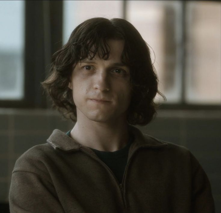 a young man with long hair wearing a brown jacket and black shirt looking at the camera