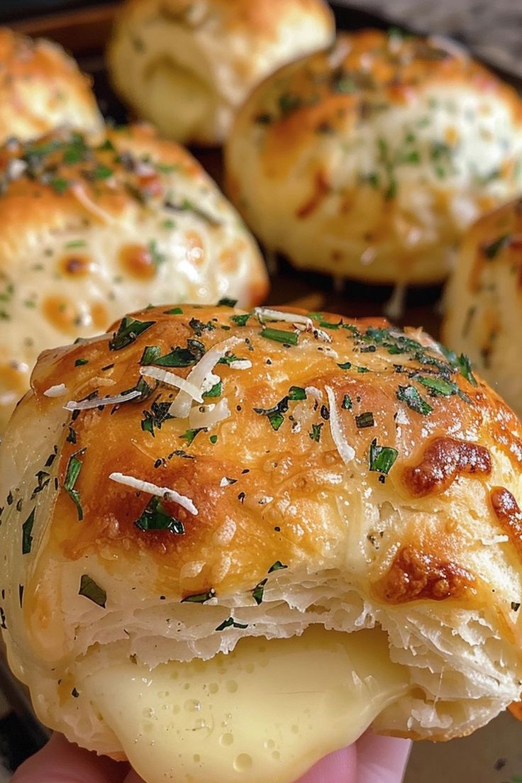 a hand holding a piece of bread with cheese and herbs on it, in front of other pastries