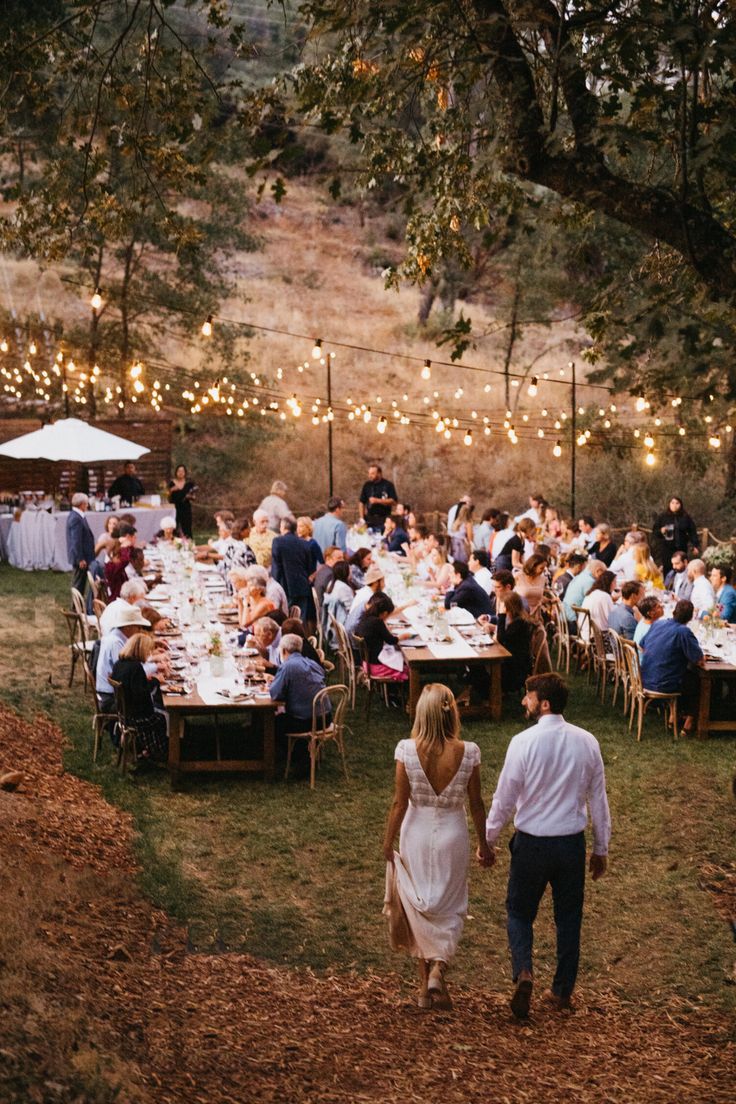 a man and woman holding hands walking through an outdoor dining area with string lights strung across the trees