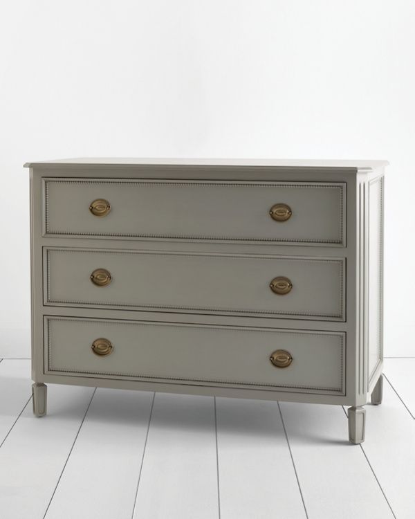 a white dresser with three drawers and gold handles on the bottom drawer, against a white wall