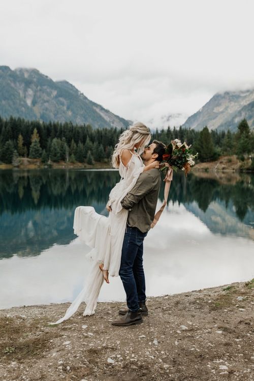 a man and woman standing next to each other near a body of water with mountains in the background