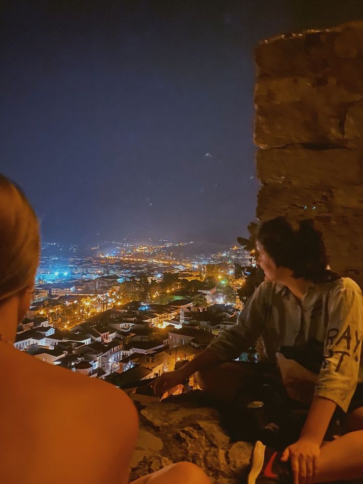 two people sitting on the edge of a cliff overlooking a city at night with lights in the distance