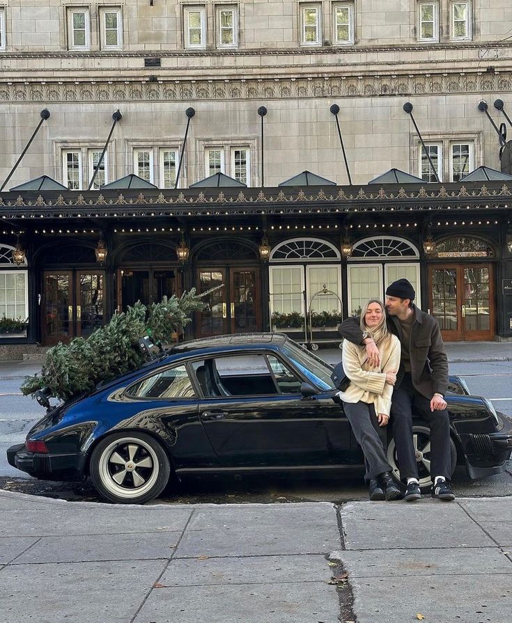 a man and woman sitting on the hood of a car in front of a hotel