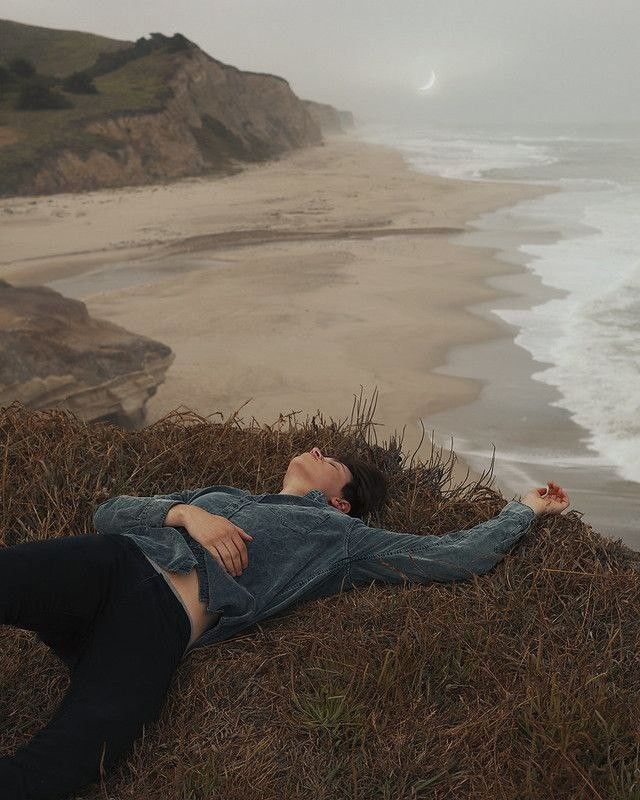 a woman laying on top of a grass covered hillside next to the ocean with her eyes closed