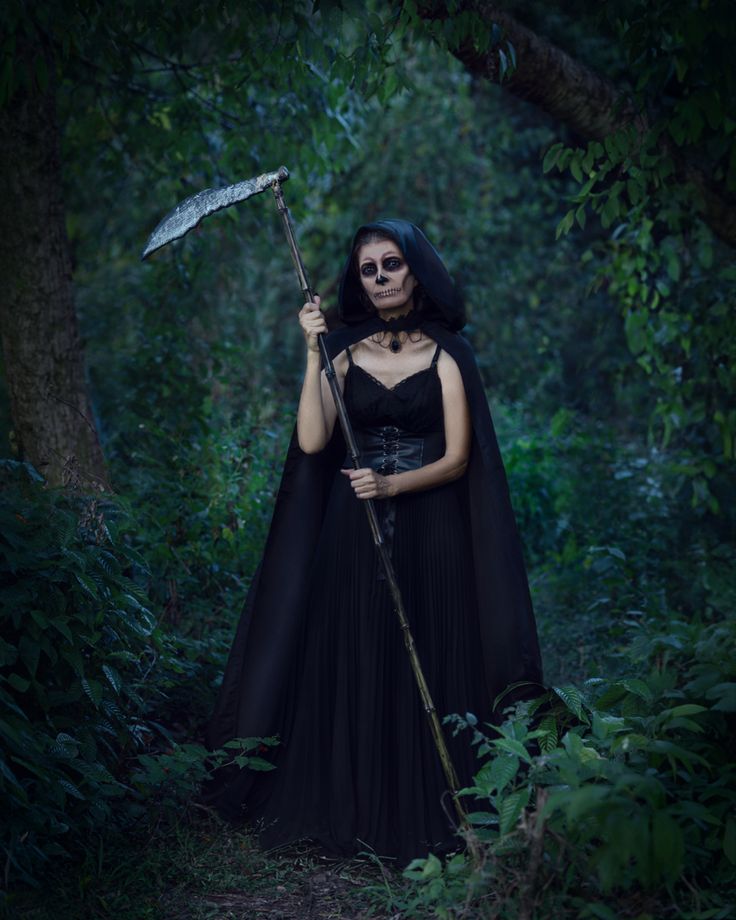 a woman dressed in black holding an umbrella and standing in the woods with her face painted like a skeleton