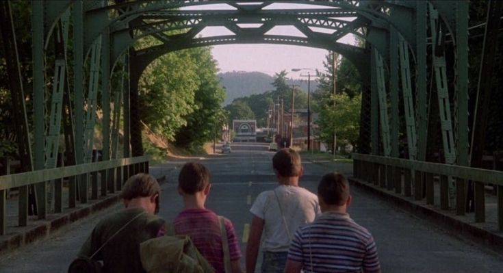 three people walking across a bridge over a road with trees on both sides and mountains in the background