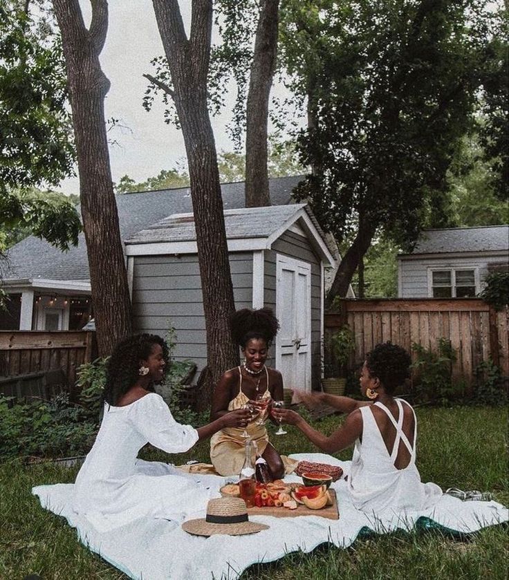 three women sitting on a blanket in the yard having a picnic and talking to each other