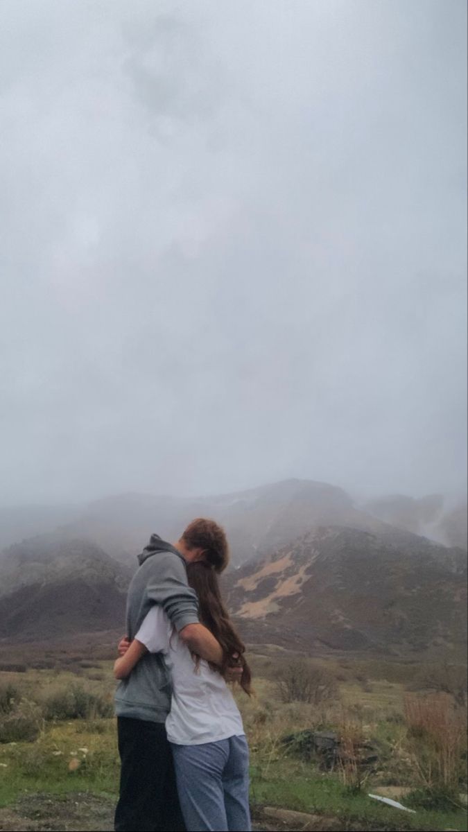 two people embracing each other in front of some hills and mountains on a cloudy day