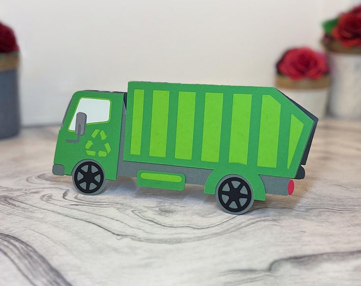 a green paper toy truck sitting on top of a wooden table next to flowers and potted plants