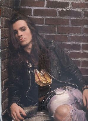 a man with long hair sitting against a brick wall wearing a leather jacket and skirt