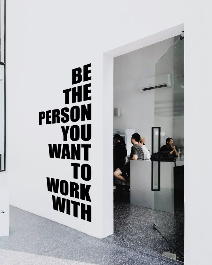 a man sitting at a desk in front of a wall with the words be the person you want to work with