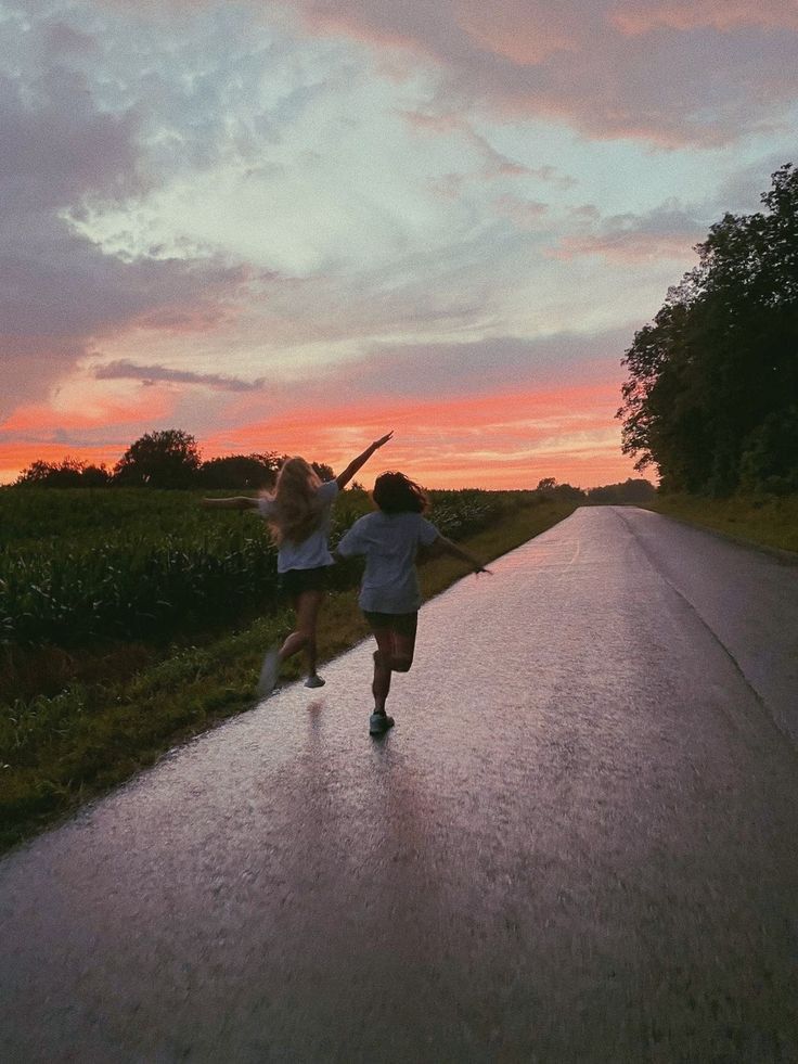 two girls are running down the road in the rain as the sun sets behind them