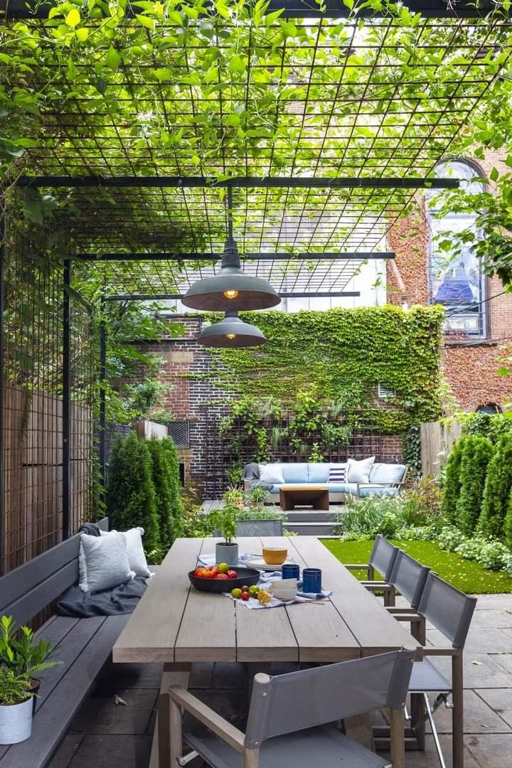 an outdoor dining area with wooden table and benches under a pergolated roof, surrounded by greenery