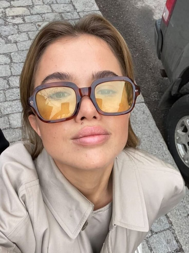 a woman wearing glasses is taking a selfie in front of a parked car and looking up at the camera