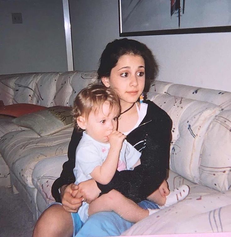 a woman sitting on top of a couch holding a baby next to her chest and looking at the camera