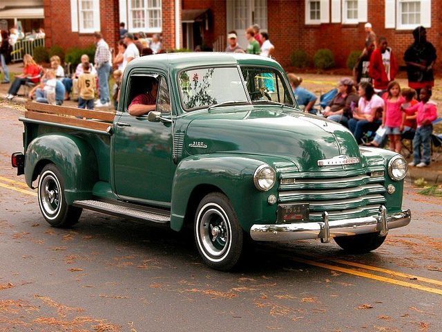 an old green truck driving down the street with people sitting on the sidelines watching