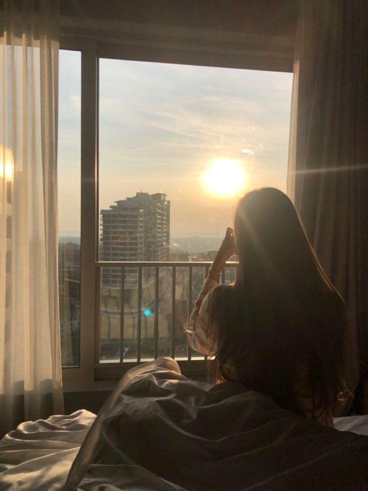 a woman is sitting in bed looking out the window at the sun setting behind her