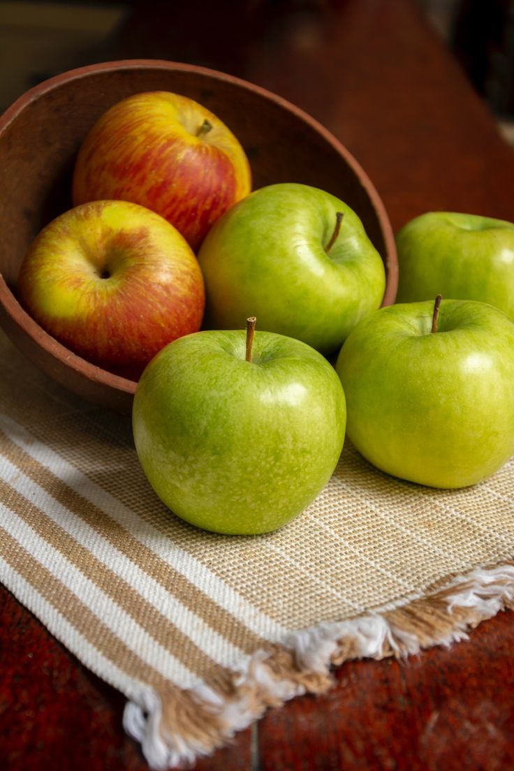 four green apples in a bowl on a tablecloth next to two red and yellow apples