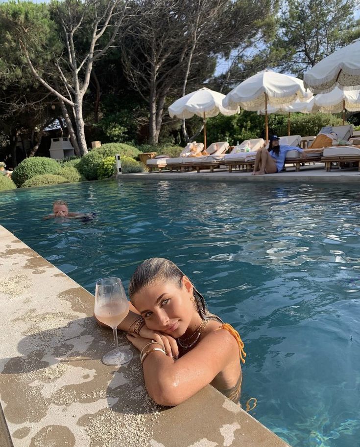 a woman leaning on the edge of a swimming pool with two wine glasses in front of her