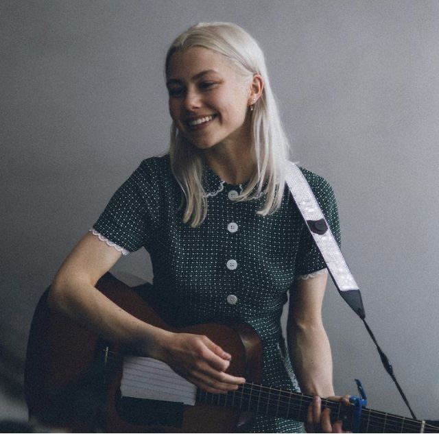 a woman with blonde hair holding a guitar and singing into a microphone while standing in front of a white wall