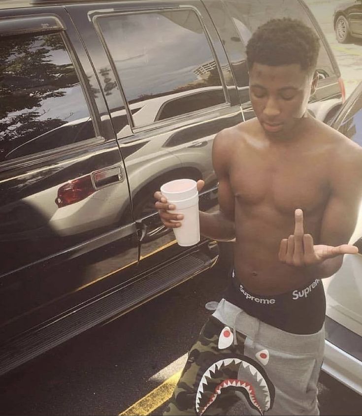 a shirtless young man holding a cup and pointing to the side while standing in front of parked cars