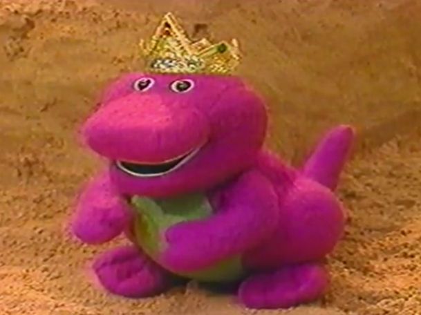 a purple toy dinosaur with a crown on it's head sitting in the sand