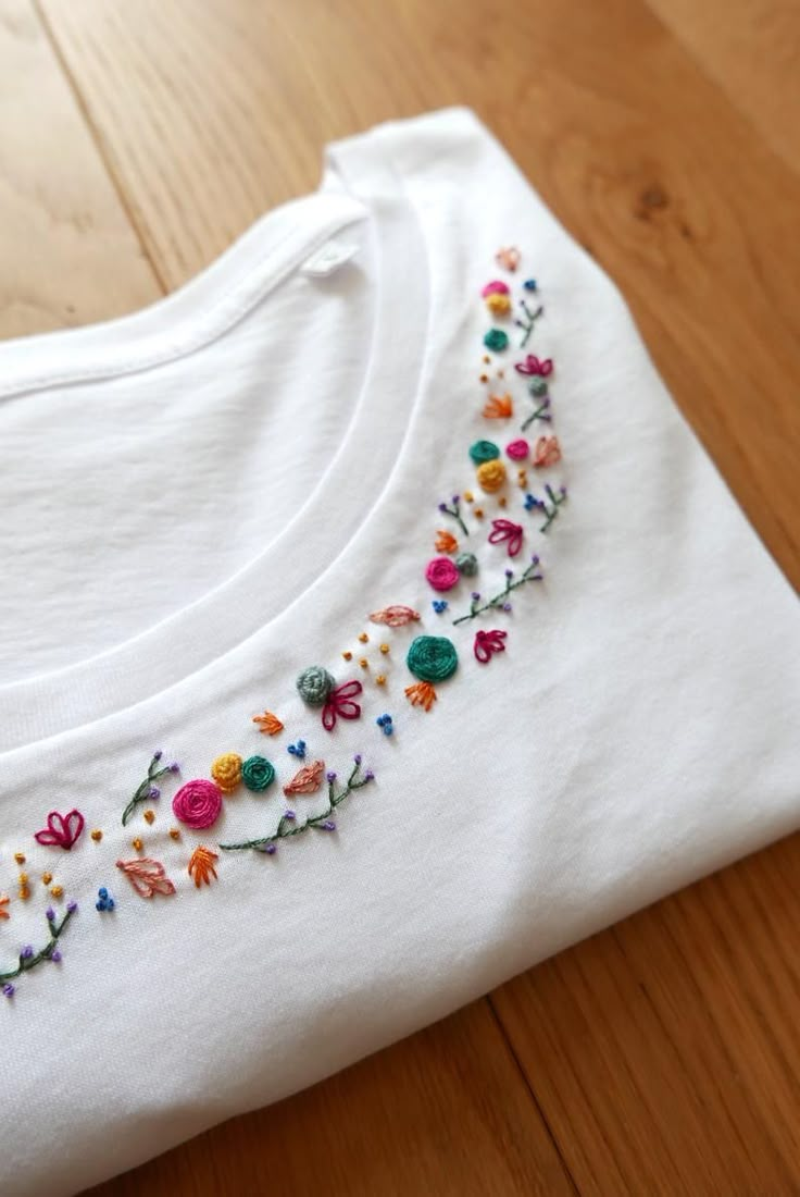 a white t - shirt with embroidered flowers on it sitting on a wooden floor next to a pair of scissors