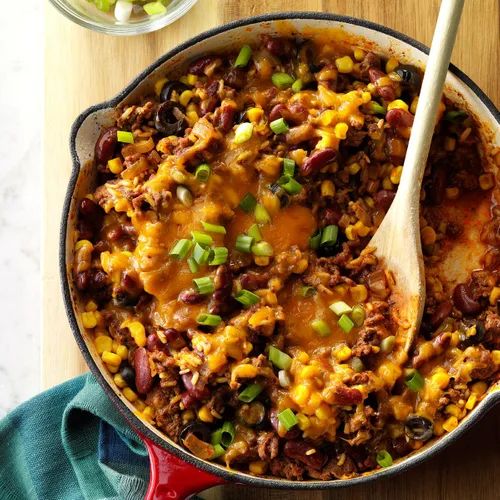 a skillet filled with chili and beans on top of a wooden table next to a bowl of green onions