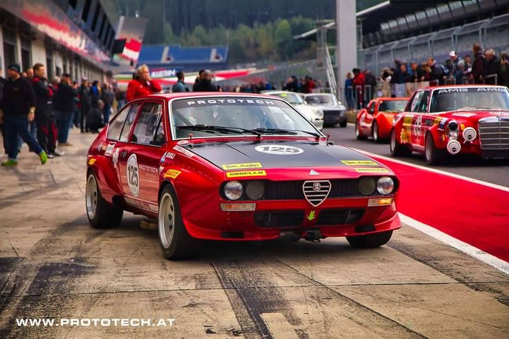 a red race car driving down a track with other cars behind it and people watching from the sidelines