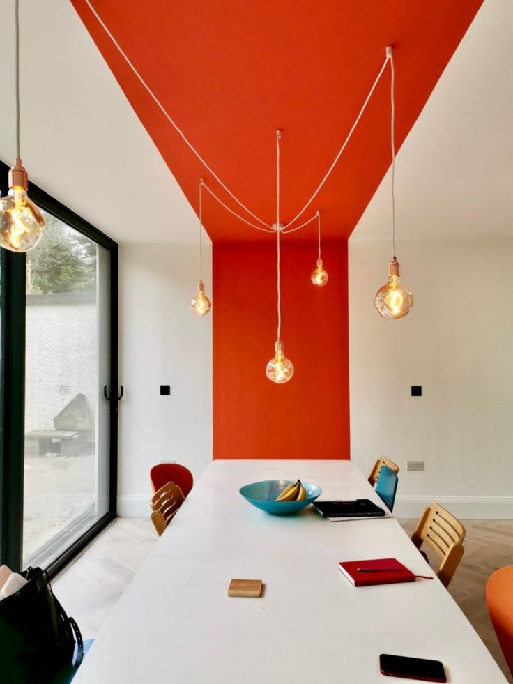 an orange and white dining room with lights hanging from the ceiling over the long table