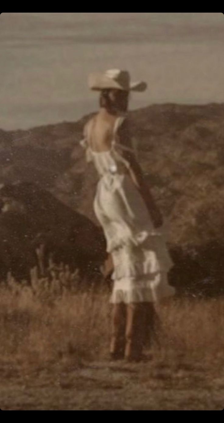 a woman wearing a white dress and hat standing in a field with mountains in the background